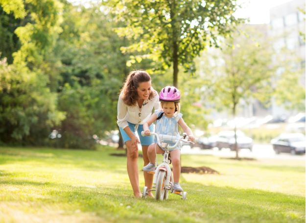 A resident of the Alta Nova apartments helps a child, wearing a pink helmet, ride a bike with training wheels in the sunny park.